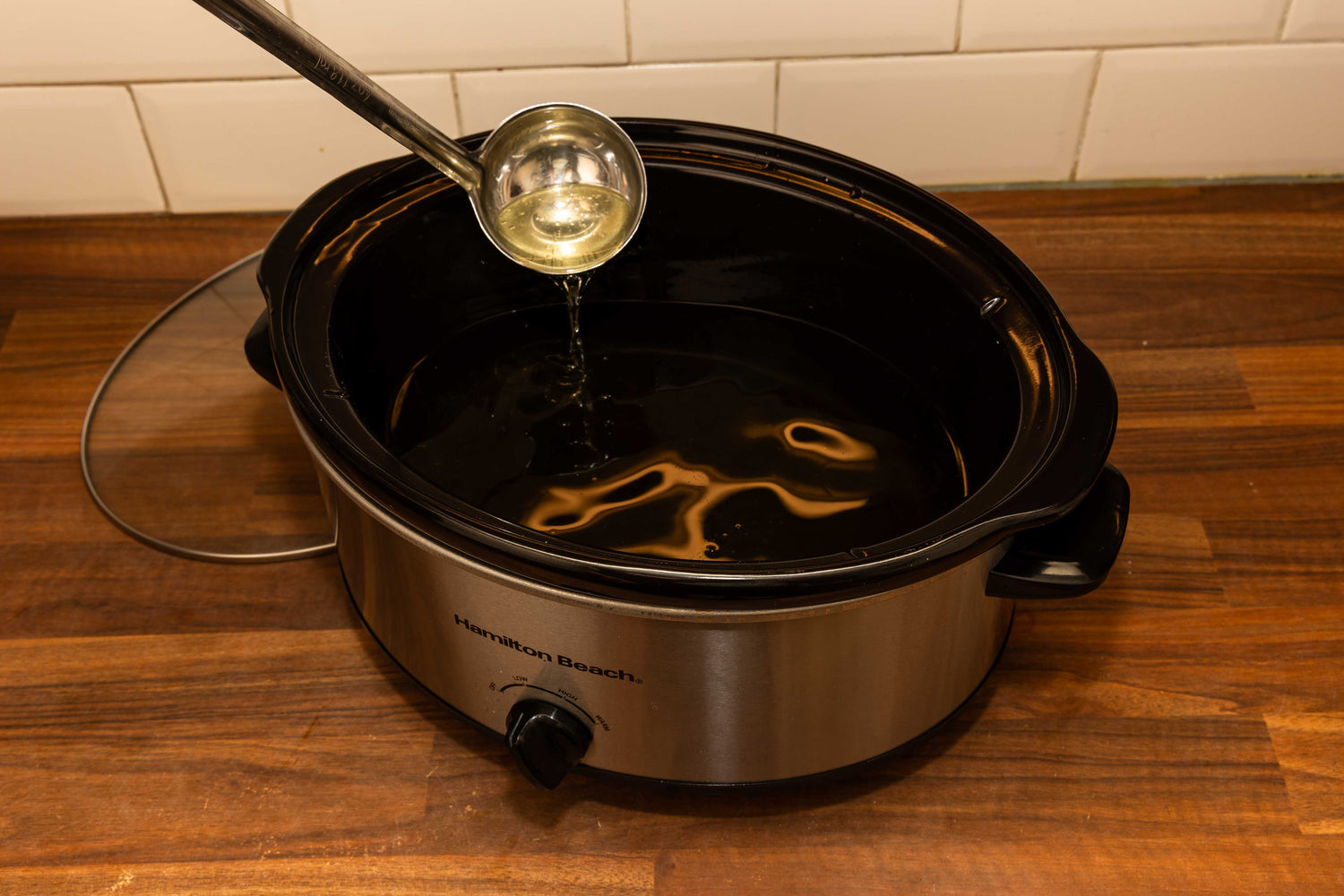 A slow cooker filled with melted tallow, with a metal ladle lifting the golden liquid. The appliance sits on a wooden countertop against a white tiled backsplash.