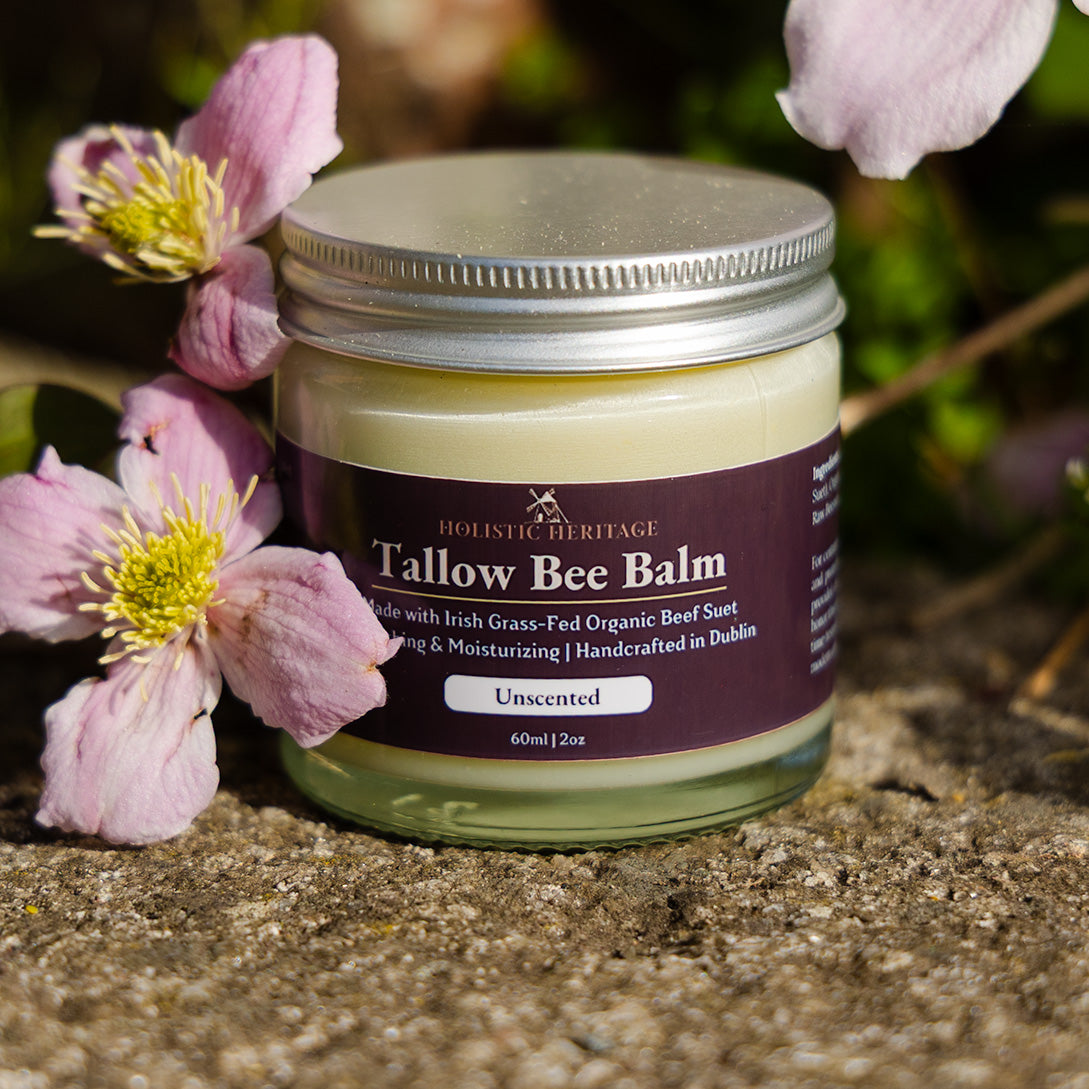Unscented Tallow Bee Balm jar on a stone surface surrounded by soft pink flowers, photographed in natural sunlight.