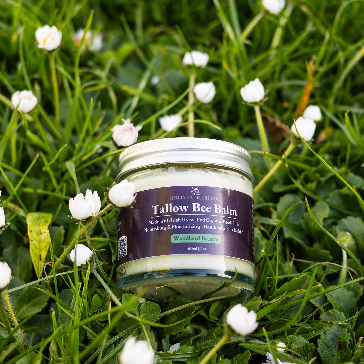 Glass jar of Holistic Heritage Tallow Bee Balm (Woodland Breathe) nestled in fresh green grass with small white flowers.