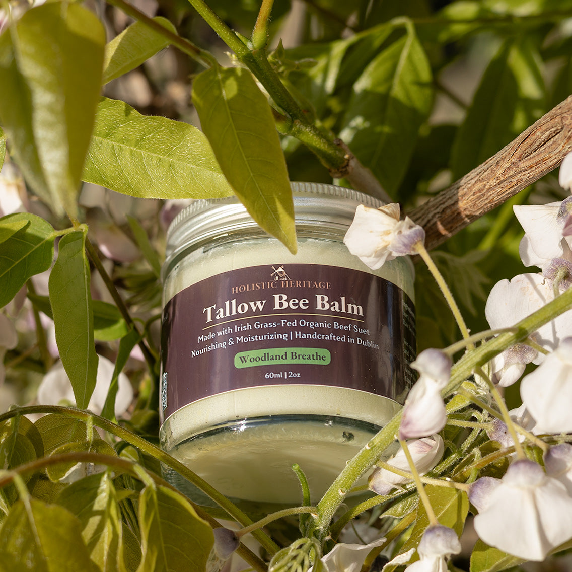 Jar of Holistic Heritage Woodland Breathe Tallow Bee Balm nestled among leafy green branches and soft white flowers in natural sunlight.